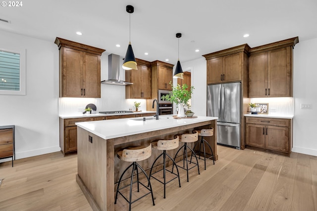 kitchen with appliances with stainless steel finishes, light hardwood / wood-style floors, an island with sink, wall chimney exhaust hood, and decorative light fixtures