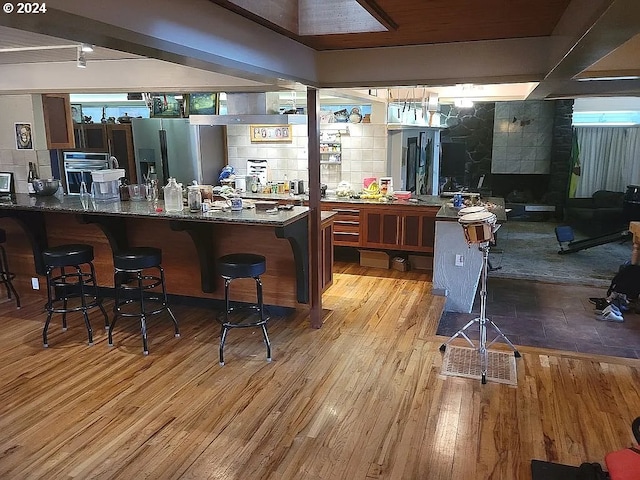 bar featuring stainless steel refrigerator with ice dispenser, light wood-type flooring, dark stone counters, and beam ceiling