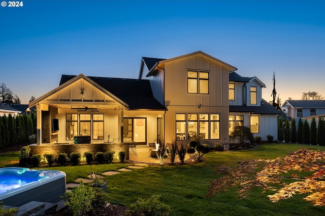 back house at dusk featuring a hot tub, ceiling fan, a patio area, and a lawn