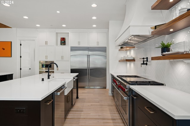 kitchen featuring premium range hood, high end appliances, sink, white cabinets, and an island with sink