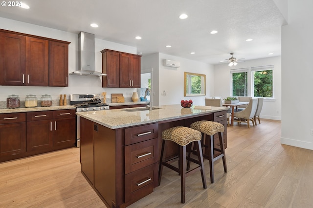 kitchen with wall chimney exhaust hood, an island with sink, light hardwood / wood-style floors, sink, and stainless steel range with gas stovetop