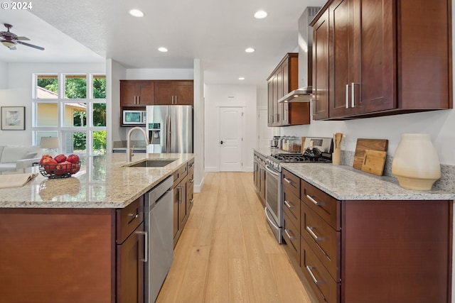 kitchen with appliances with stainless steel finishes, wall chimney exhaust hood, sink, light stone counters, and light hardwood / wood-style flooring