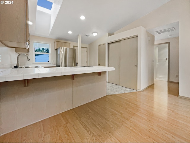 kitchen with sink, stainless steel fridge, light hardwood / wood-style floors, vaulted ceiling with skylight, and tile counters