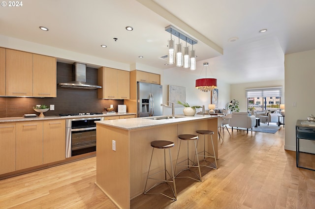 kitchen with appliances with stainless steel finishes, wall chimney exhaust hood, light brown cabinets, and a center island with sink