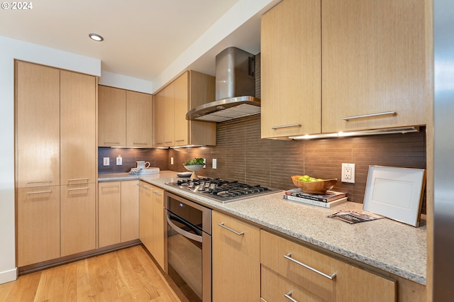 kitchen featuring tasteful backsplash, wall chimney range hood, light stone countertops, appliances with stainless steel finishes, and light brown cabinetry