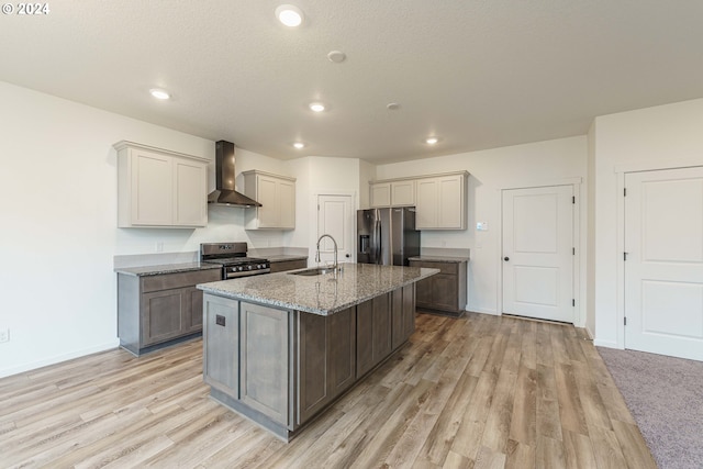 kitchen with sink, wall chimney exhaust hood, stainless steel appliances, an island with sink, and light hardwood / wood-style floors