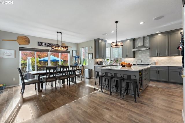 kitchen featuring gray cabinetry, wall chimney exhaust hood, dark wood-type flooring, an island with sink, and pendant lighting