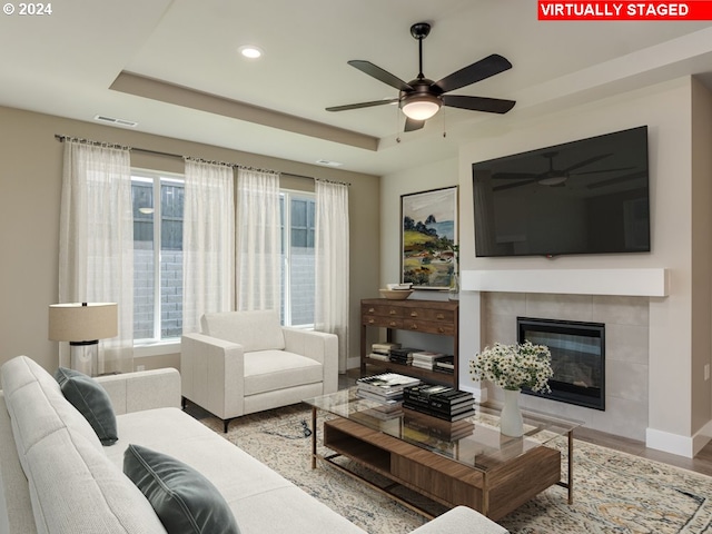 living room featuring a tray ceiling, wood-type flooring, a tile fireplace, and ceiling fan