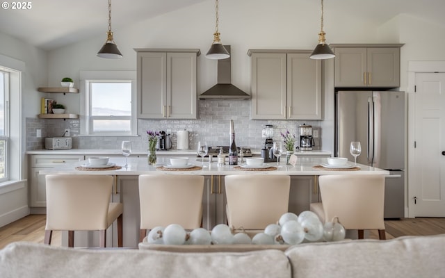 kitchen featuring stainless steel refrigerator, an island with sink, vaulted ceiling, and wall chimney range hood