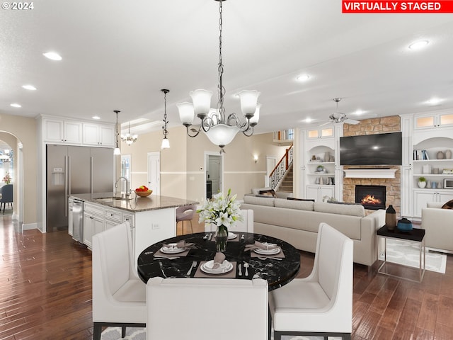 dining room with arched walkways, a stone fireplace, built in shelves, dark wood-type flooring, and a ceiling fan