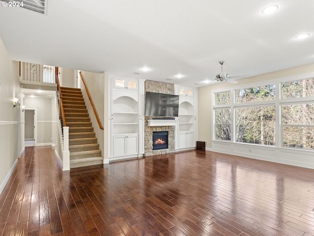 unfurnished living room featuring built in shelves, a fireplace, dark wood finished floors, stairway, and ceiling fan