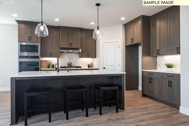 kitchen with built in microwave, stainless steel oven, decorative light fixtures, and an island with sink