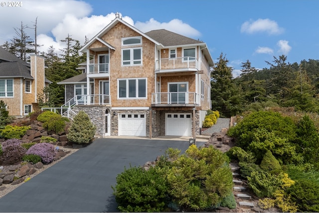 view of front of house with a shingled roof, an attached garage, a balcony, stone siding, and driveway