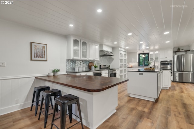 kitchen with appliances with stainless steel finishes, white cabinetry, wall chimney exhaust hood, a kitchen island, and light hardwood / wood-style flooring