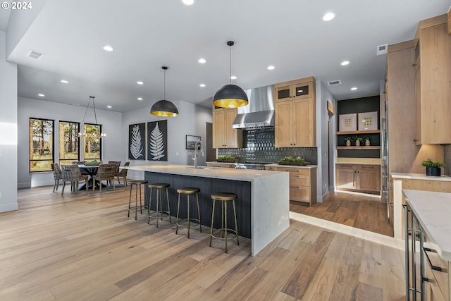 kitchen featuring a center island with sink, light hardwood / wood-style flooring, hanging light fixtures, and wall chimney range hood