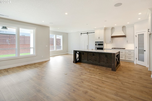 kitchen featuring light hardwood / wood-style floors, tasteful backsplash, a kitchen island with sink, a barn door, and premium range hood