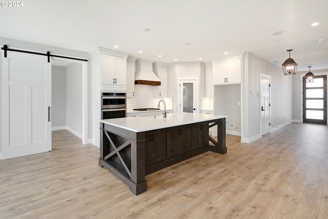 kitchen with light wood-type flooring, a center island with sink, stainless steel double oven, premium range hood, and a barn door