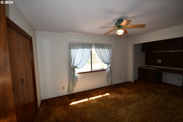 unfurnished bedroom featuring ceiling fan, built in desk, dark carpet, and a closet