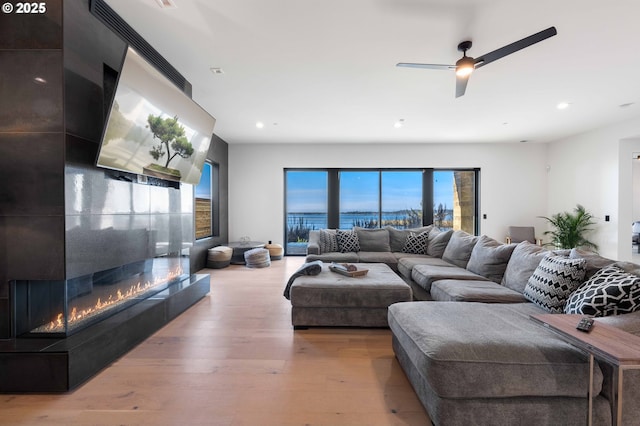 living area with a glass covered fireplace, light wood-style flooring, recessed lighting, and ceiling fan