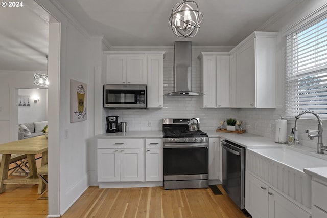 kitchen with light wood-type flooring, white cabinetry, wall chimney range hood, stainless steel appliances, and decorative light fixtures