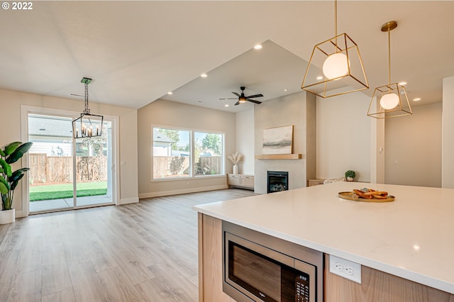 kitchen with light wood-type flooring, hanging light fixtures, stainless steel microwave, and ceiling fan with notable chandelier