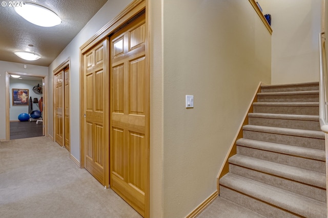 hallway featuring light colored carpet, a textured ceiling, baseboards, and stairs