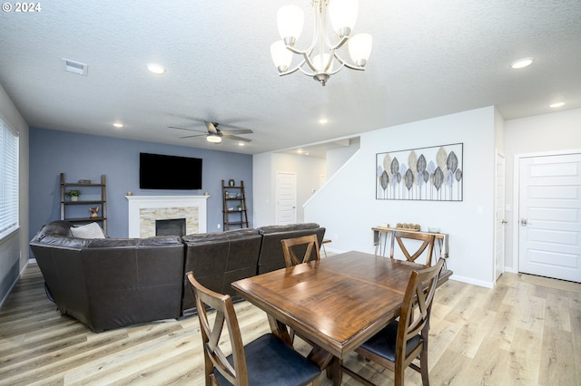 dining area with ceiling fan with notable chandelier, light hardwood / wood-style floors, a stone fireplace, and a textured ceiling