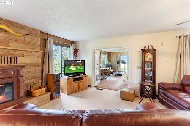 living room featuring ceiling fan, a stone fireplace, a textured ceiling, wooden walls, and light carpet