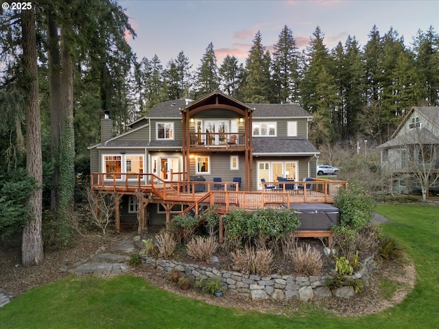 back house at dusk featuring a hot tub, a wooden deck, a balcony, and a lawn