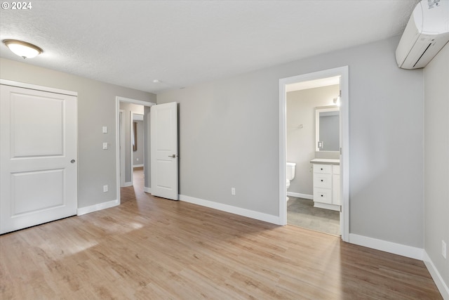 unfurnished bedroom featuring ensuite bathroom, a textured ceiling, a wall unit AC, and light hardwood / wood-style flooring