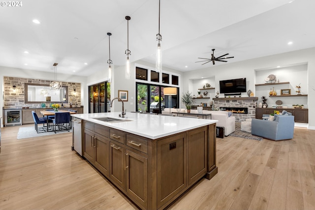 kitchen with wine cooler, a stone fireplace, sink, a center island with sink, and pendant lighting