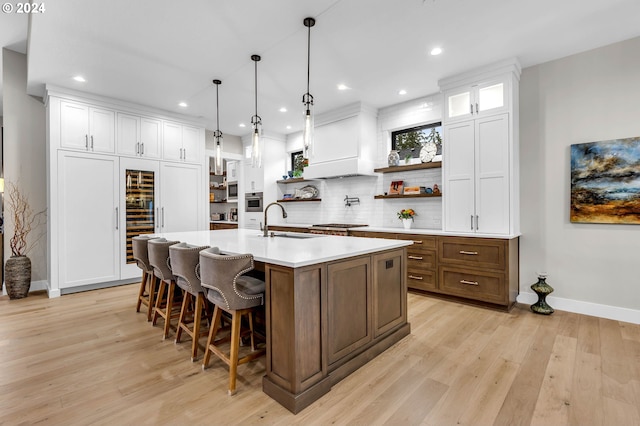 kitchen with decorative light fixtures, an island with sink, sink, white cabinets, and light wood-type flooring