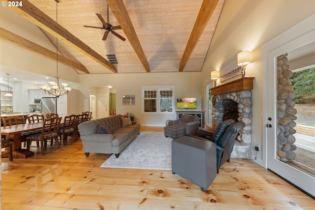 living room with ceiling fan with notable chandelier, light wood-type flooring, beamed ceiling, and a stone fireplace