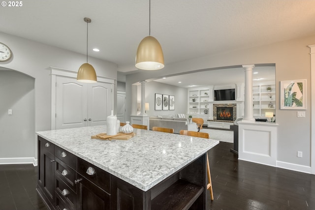 kitchen featuring built in shelves, dark wood-type flooring, pendant lighting, a center island, and a stone fireplace