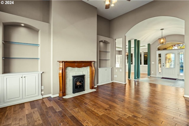 unfurnished living room featuring built in shelves, a high end fireplace, hardwood / wood-style floors, a towering ceiling, and ceiling fan with notable chandelier