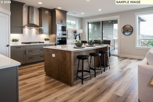 kitchen featuring a wealth of natural light, stainless steel appliances, wall chimney range hood, hardwood / wood-style floors, and a kitchen island with sink