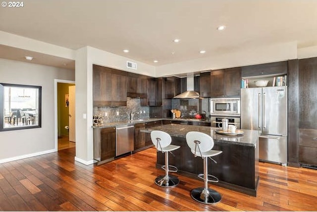 kitchen featuring a center island, wall chimney exhaust hood, stainless steel appliances, decorative backsplash, and hardwood / wood-style flooring