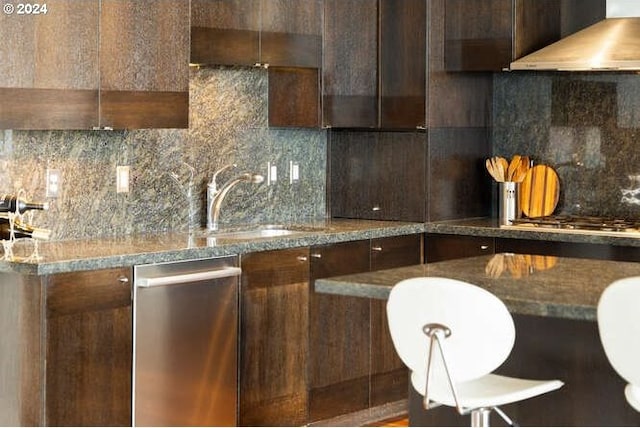 kitchen with dark stone countertops, sink, wall chimney range hood, and backsplash