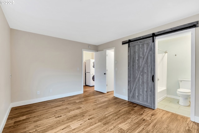 unfurnished bedroom with stacked washer and clothes dryer, a barn door, light wood-type flooring, and ensuite bath