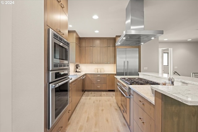 kitchen featuring built in appliances, backsplash, light hardwood / wood-style flooring, island range hood, and light stone counters