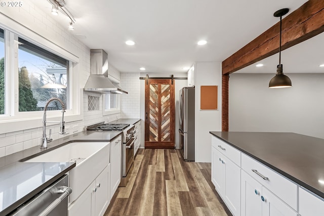 kitchen featuring appliances with stainless steel finishes, wall chimney exhaust hood, pendant lighting, a barn door, and white cabinetry