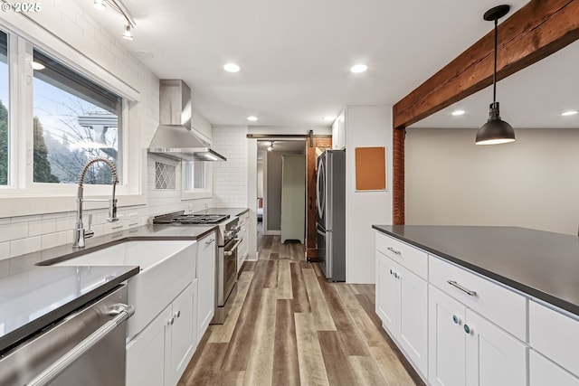 kitchen featuring white cabinets, wall chimney exhaust hood, a barn door, appliances with stainless steel finishes, and decorative light fixtures