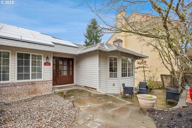 entrance to property with a patio, roof mounted solar panels, a shingled roof, brick siding, and a chimney