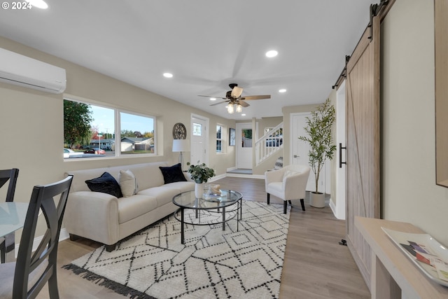 living room with a wall mounted AC, a barn door, light wood-type flooring, and ceiling fan