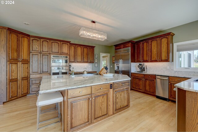 kitchen with an island with sink, hanging light fixtures, light hardwood / wood-style flooring, stainless steel appliances, and decorative backsplash