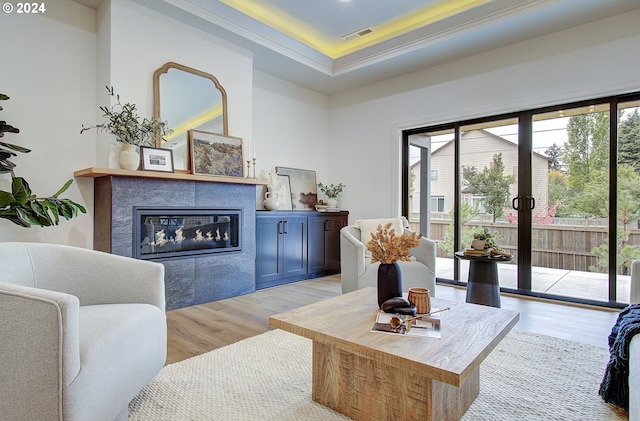 living room featuring visible vents, a raised ceiling, a glass covered fireplace, crown molding, and light wood-type flooring