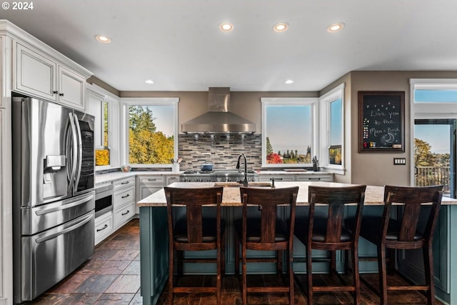 kitchen with an island with sink, appliances with stainless steel finishes, decorative backsplash, light stone countertops, and wall chimney range hood