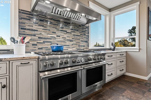 kitchen featuring backsplash, wall chimney range hood, range with two ovens, white cabinets, and light stone counters