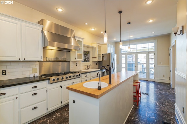 kitchen featuring stainless steel appliances, white cabinetry, a kitchen island with sink, wall chimney exhaust hood, and butcher block counters