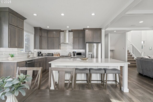 kitchen featuring wall chimney exhaust hood, a kitchen bar, a center island, appliances with stainless steel finishes, and dark hardwood / wood-style flooring